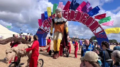 BBC Scene at Eisteddfod with arch spelling out Eisteddfod and 850 either side to mark the festival's anniversary. A very large puppet is visible under the arch with people in red jumpsuits behind moving it along with the help of wheels as people look on. A smaller dog puppet on wheels is visible in front.