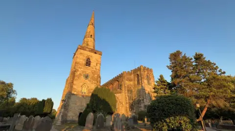 A church surrounded by trees and a graveyard. There is a square tower with a conical spire and a large adjoining building with stained glass windows. The building is set against a blue sky.