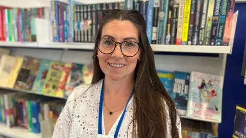 Michelle Lord has long dark hair, she wears a white top and round dark glasses. She stands in front of a bookshelf which is full of colourful books.