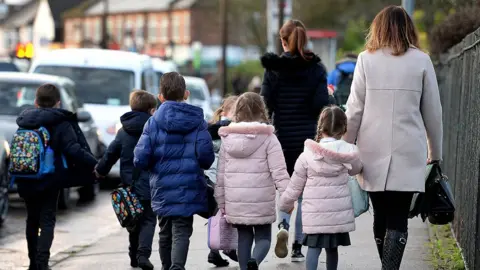 PA Media A group of young pupils and two parents walk along a pavement facing away from the camera. Two of the girls are wearing identical pink coats, while three boys where dark coats with backpacks