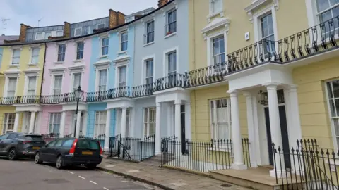 BBC A row of colorful houses on Chalcot Crescent, Primrose Hill, London. The houses have four or five floors each and are yellow, pink, blue and gray. There are two black cars parked outside.