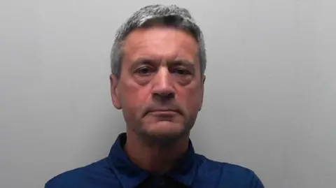 A man faces the camera against a plain light-grey background. He has short grey hair and is wearing a dark blue collared shirt.