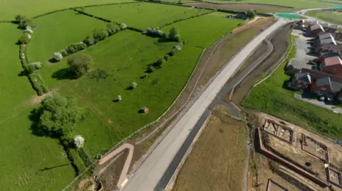 Green fields and trees to the left of a new distributor road in Melton, Leicestershire, and houses and construction work to the right.