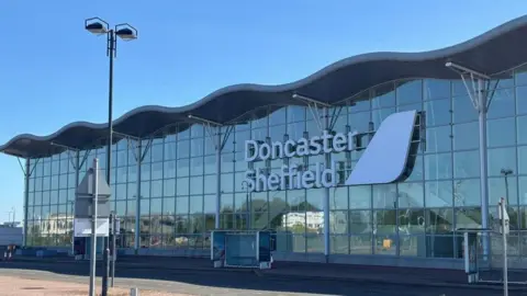 An image of the Doncaster Sheffield Airport terminal. It is a large glass-fronted building with a wavy shaped roof. There is a large white sign saying Doncaster Sheffield.