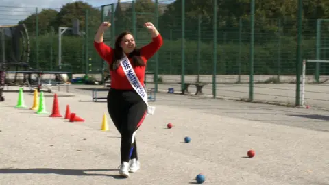 Oliver Whitfield-Miočić/BBC A woman with a red top and black sports leggings walks with her hands in the air on a sports field. 