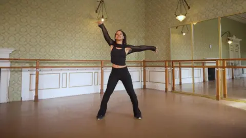 Particle6 Anais Houillet performing a dance move in a room, with mirrors. She is wearing an all black dance outfit.