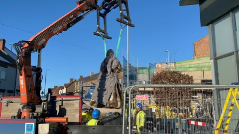 Workmen use a crane to move a large statue towards a plinth