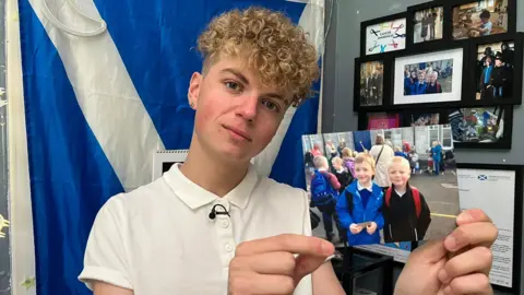 Ryan Quinn, a blonde, curly-haired boy, looks at the camera while holding a photograph of a young Aaron and Ryan at school. On the wall behind him are hung a selection of his other favourite photos of the pair, and a Scotland flag is pinned up on the adjacent wall.
