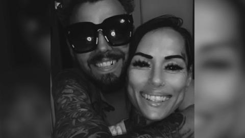 Ben Atkins and Debbie Pereira smiling at the camera in this black and white image. He is wearing dark sunglasses and is hugging her