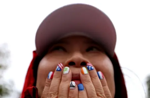  Steph Chambers/Getty Images A woman wearing a cap covers her mouth with her hands. Her nails are painted with flags and symbols