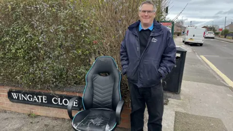 A man with grey hair and a navy blue coat stands at the end of a residential street corner, signed 'Wingate Close'. He is stood with his hands in his pockets next to a worn down black leather office chair on the pavement.
