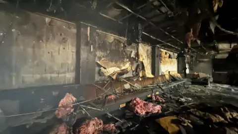 The interior of the burned-out shell of the dance school in Clubmoor, Liverpool. Beams are exposed in the ceiling, with insulation lying around on the floor and blackened debris everywhere.