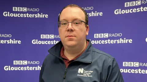 Stewart looks at the camera. He has wire rimmed glasses and short brown hair. He is wearing a navy zipped jacket with an RNC logo on the label. Behind him is the BBC Radio Gloucestershire purple backing.