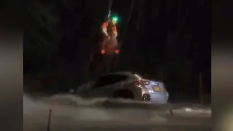 Two people are airlifted from above a car half-submerged in water at night.