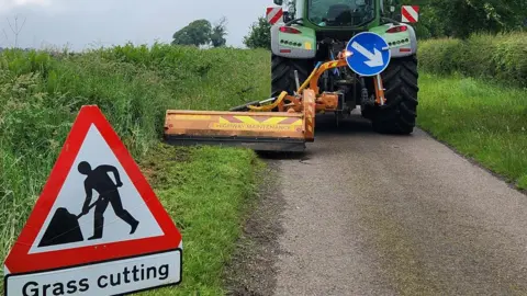 A red road sign reads 'grass cutting' and has a person digging into the ground. The sign is place behind a tractor which has a grass cutting strimmer fixed to its left side. It is a small single-track road which has had its verge partly cut. 