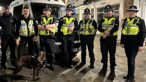 A seven-member police team, including officers dressed in black uniforms and hi-vis vests, stand in front of a white police van with a brown English pointer dog, in a pedestrian area of Beverley town centre.