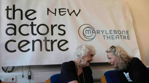 Gavin Watson Two women on blue chairs lean in to chat in front of a sign for The New Actors Centre at Marylebone Theatre