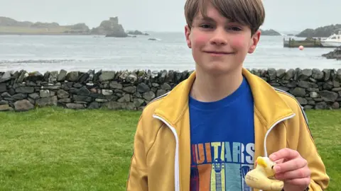 Emil Wrede, a young boy, holds up a plastic duck, while smiling. He has short, brown hair and is wearing a blue T-shirt with a yellow jacket