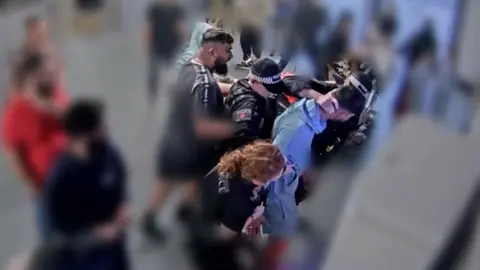 Screenshot from CCTV footafe of two men in an altercation with three police officers. People surrounding them are blurred. 