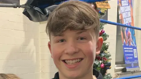 Charlie has brown hair and is in a corridor with white painted bricks.