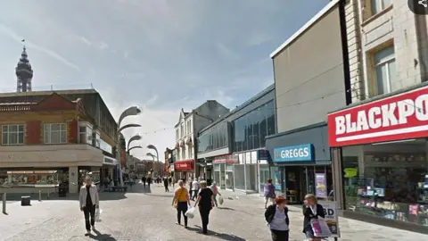 Google Shoppers walking on Church Street in Blackpool town centre. Blackpool Tower is in the background. The photograph was taken on a sunny day.