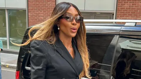 PA Media Naomi Campbell has long blonde and brown straight hair, brown sunglasses and a black blazer jacket. She is walking along, having just got out of a black car.