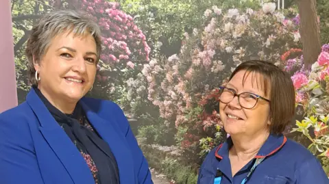 Bradford Teaching Hospitals Two women are stood in front of a wall with images of flowers on it. The woman on the left is wearing a royal blue blazer and a navy blue scarf, she has short, light grey hair and hooped earrings. The woman on the left has a brown bob and is wearing glasses, she is wearing a navy blue nurses gown. Both are smiling.
