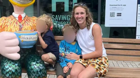 Astrid Cooper Astrid Cooper wearing a white vest and shorts with a sunflower pattern. She is sitting on a wooden bench beside her two young children, who have their faces blurred. 