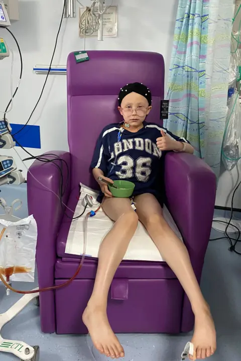 Danielle Roberts A 10 year old girl sits in a purple hospital armchair. She is wearing pyjama shorts and a t-shirt with a baseball style logo that reads London 36. She is wearing a black head scarf with white polka dots and glasses and is attached to medical machinery with tubes. She looks sad but is making a thumbs up with one hand and holding a green bowl in the other.