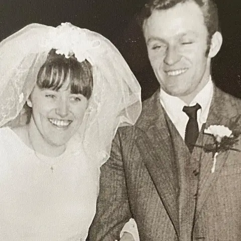 Jackie Clifton A black and white photograph of a couple at their wedding. Both are smiling. The bride, on the left, is wearing a white dress and a small crucifix necklace. Her veil is off her face and has a floral decoration at the top. Her husband is wearing a suit, a white shirt and dark tie. He has a carnation flower pinned to his jacket lapel.