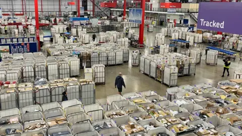 Large crates of post being prepared for delivery at the Medway Mail Centre in Kent; personnel can be seen moving around the centre floor, where there are rows and rows of metal crates with white bags. On the right hand side there is a section for tracked mail.