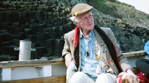 PA Media Peter Monro. He is an elderly man sitting on a boat in front of some cliffs. He is wearing a flat cap, a blue shirt and a plaid blazer featuring various patches of colorful fabric, and a red velvet scarf. He is looking to the side away from the camera.