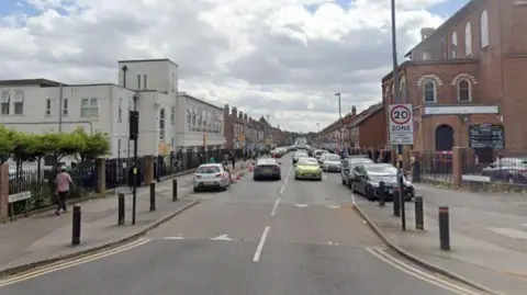 A built-up street with a 20mph zone sign and buildings on either side. Cars drive down on both sides and there are also cars parked on the side.