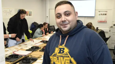 Hull City Council A man with short brown hair is wearing a blue hoodie with yellow writing on it and smiling into the camera with stubble. Behind him is a table full of people looking at bits of paper and laptops, headsets and mice devices can also be seen.