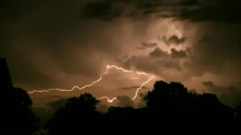 Weather Watchers/Dotty A large lightning bolt over the sky in Swindon. The sky has an orange tone from the flash. There are dark clouds and silhouettes of trees also in the frame.