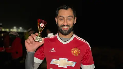 Ismail smiles at the camera as he holds the trophy after the race. He's wearing a Manchester United football top with a Jersey Spartan AC race number on it. He as a grey lycra top under his shirt. He has a short dark beard and short dark hair. Runners are standing behind him in the dark.