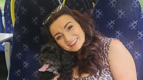 Joanne Rankin A woman with reddish/brown hair wearing a leopard print top and sunglasses on her head. She is cuddled into a black dog with her
