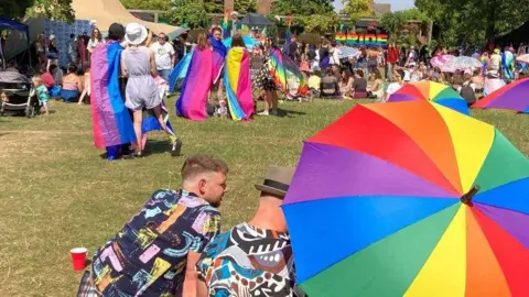 Two men wearing patterned shirts are sitting on the grass in front of a rainbow-coloured umbrella. One is wearing a green trilby hat. In front of them is a crowd of people in colourful outfits, some with Pride flags, drinking and taking in the sunshine. There are some people sitting on blankets and some children in prams. In the background is a sign which says "PRIDE" in rainbow lettering.