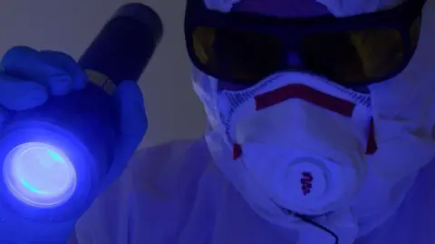 Jim Gildea is shown in full PPE, holding a torch emitting a blue light. He wears tinted lenses over his protective glasses as he looks for evidence of any remaining biological matter.