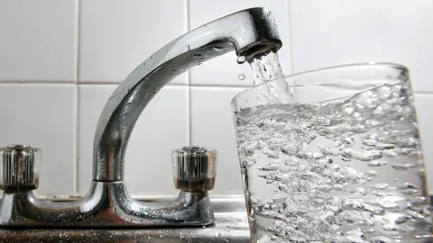 A glass of water is being filled from a silver tap. Water can be seen coming out of the spout and filling up a clear glass.