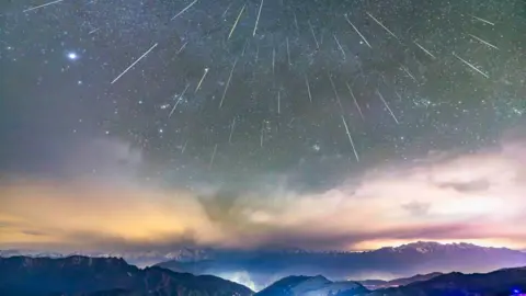 Getty Images Beberapa garis meteor terang dari hujan meteor Geminid melintas di langit penuh bintang di atas pegunungan berlapis, dengan jalan-jalan terang dan lampu-lampu terlihat di lereng Gunung Niubei di Ya'an, Provinsi Sichuan, Tiongkok.