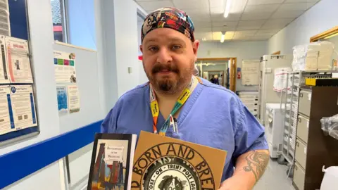 A man wearing blue medical scrubs is standing in a hospital office. There are filing cabinets behind him and various notices on a noticeboard. The man is holding a book and a commemorative plate.