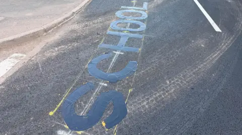 A newly laid road surface with lettering that has been blacked-out. The word "school" can be made out.