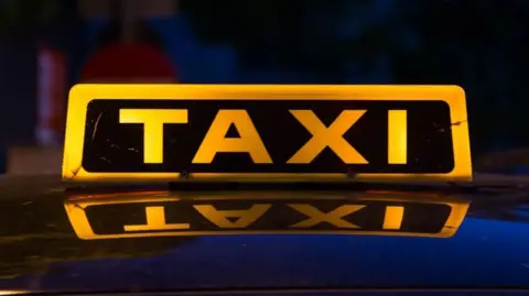 Getty Images A close up sign in bright yellow lit up saying TAXI, with the sign reflected in the car's roof.