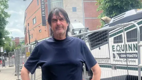a man with longish dark hair wear a dark blue tshirt. he is standing in a street in front of security barriers, with a large shopping centre in the background