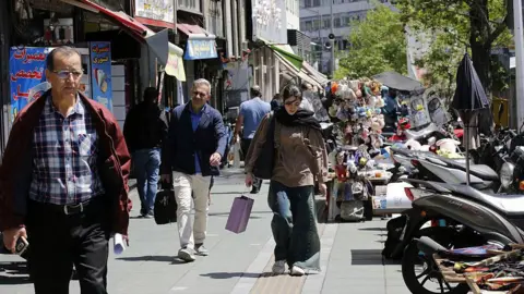 People walking on a busy shopping street in Tehran, with a young woman carrying a shopping bag in the centre of the frame