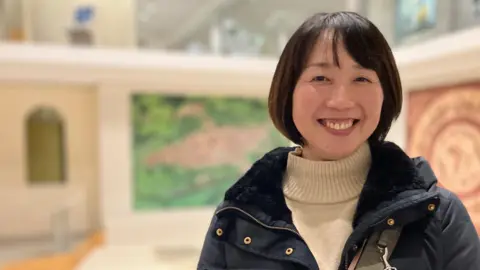 A Japanese woman is smiling and looking directly at the camera. She is wearing a navy blue coat and cream roll neck jumper. She has dark hair cut in a bob.