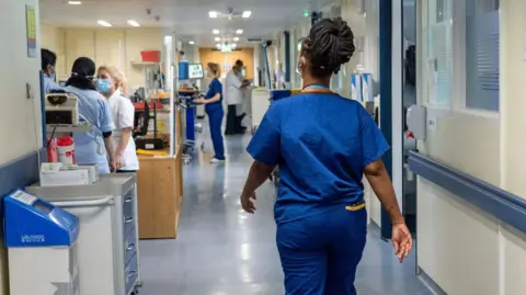 A medic walks down a corridor in a hospital. She has her back to the camera so her face cannot be seen. She is wearing blue scrubs and she has black dreadlocks tied up in a bun. Other medics wearing masks can be seen further along the corridor.