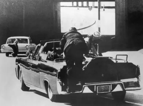 Getty Images A black and white photo of a man perched on the back of a limousine, Mrs Kennedy also with her back to the camera slightly to the right