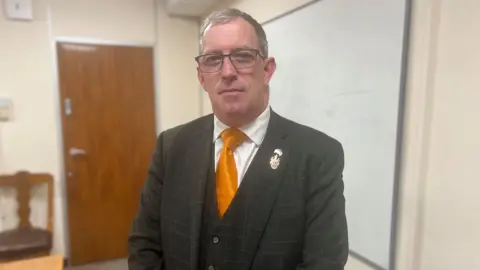 Man with glasses in chequered black suit, waistcoat, orange tie, white shirt stood infront of white board 